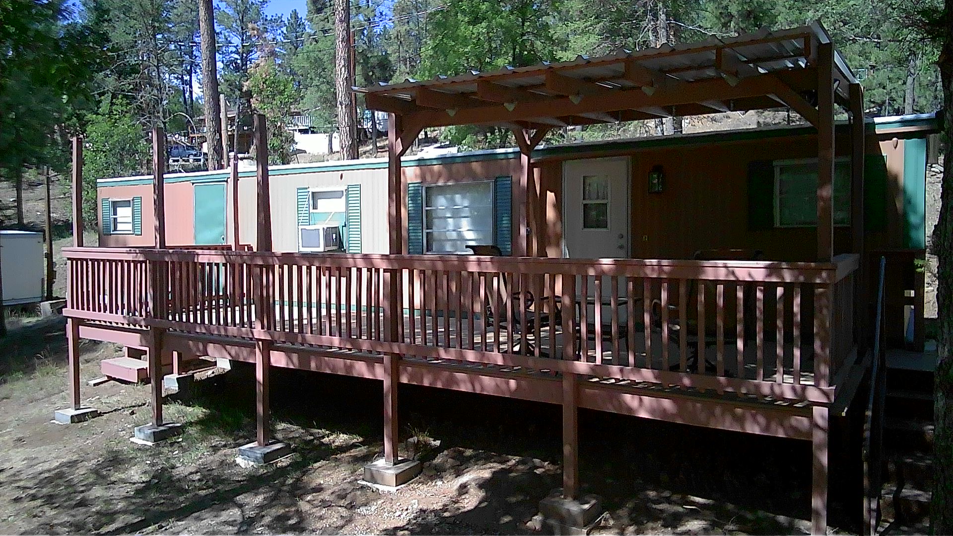 Side profile of the cabin displaying the wooden deck, roof structure with porch overhang, and surrounding tall pine trees in a serene woodland environment creating a tranquil atmosphere